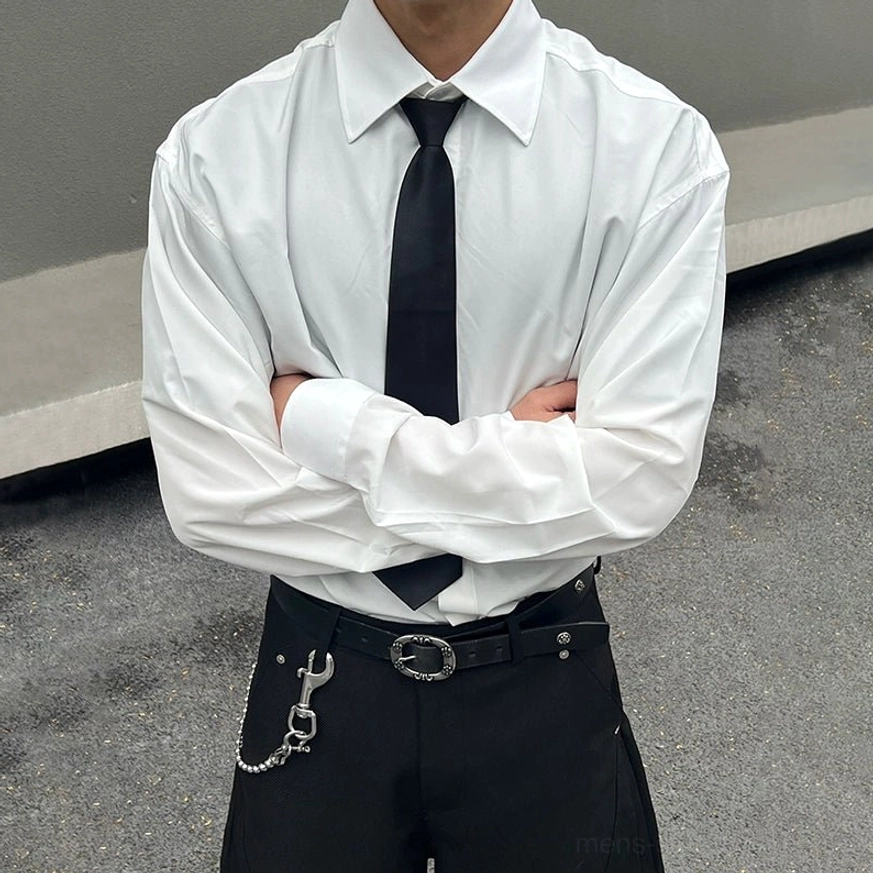 Cropped front view of an oversized white pointed collar dress shirt styled with a black tie, wide-leg trousers, chain belt, and silver stud details in a modern streetwear outfit