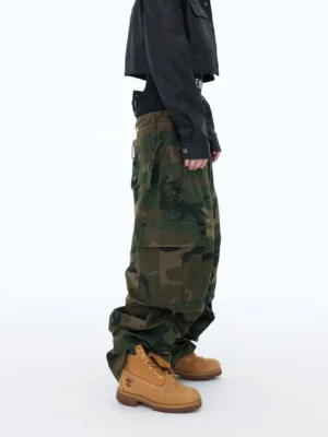 Left side view of oversized woodland camo cargo pants showing large flap pocket, loose wide-leg silhouette, drawstring waist, and stacked hem over work boots