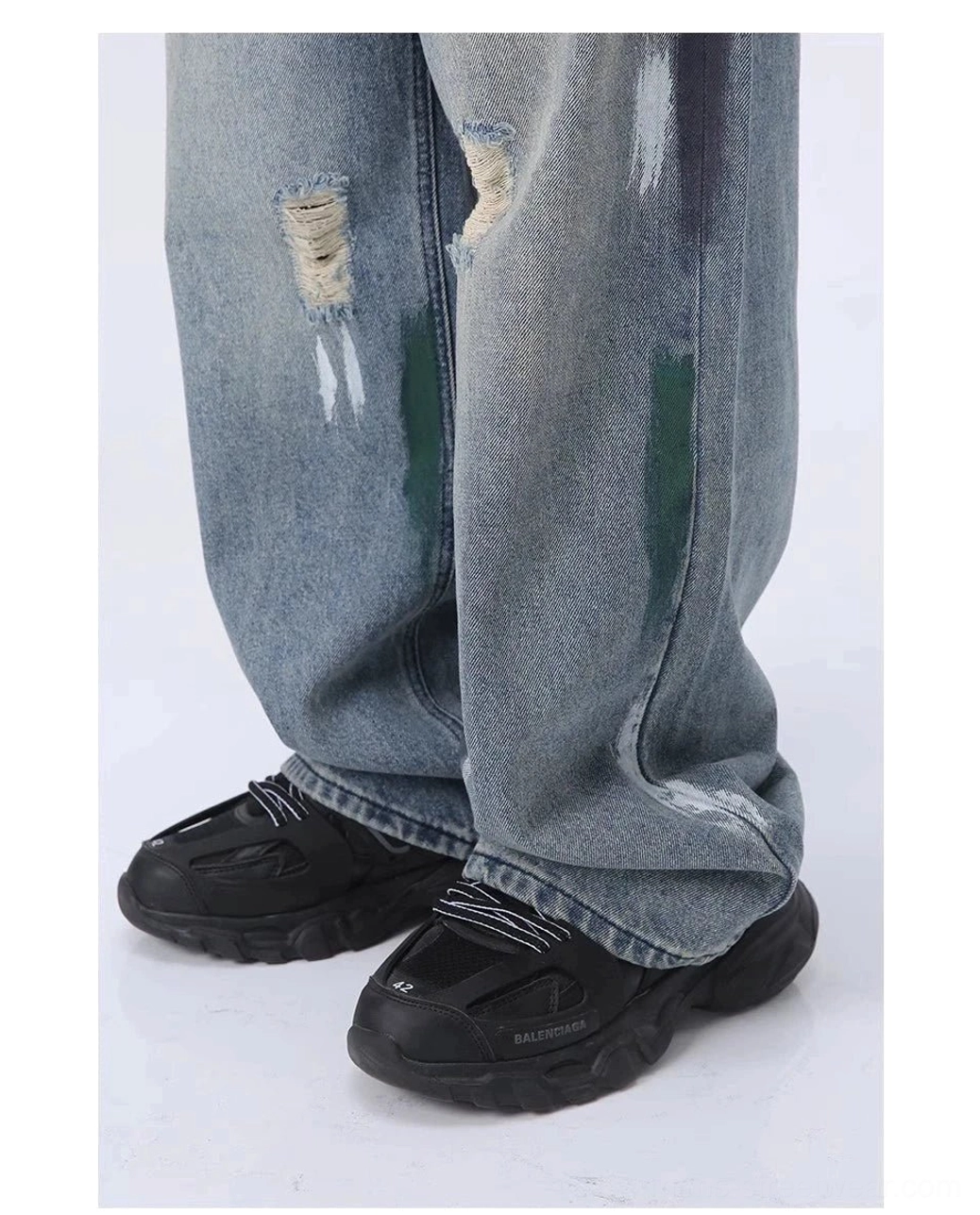 Close-up of frayed ripped lower legs and green paint splatter on light blue wide leg distressed jeans worn with chunky sneakers