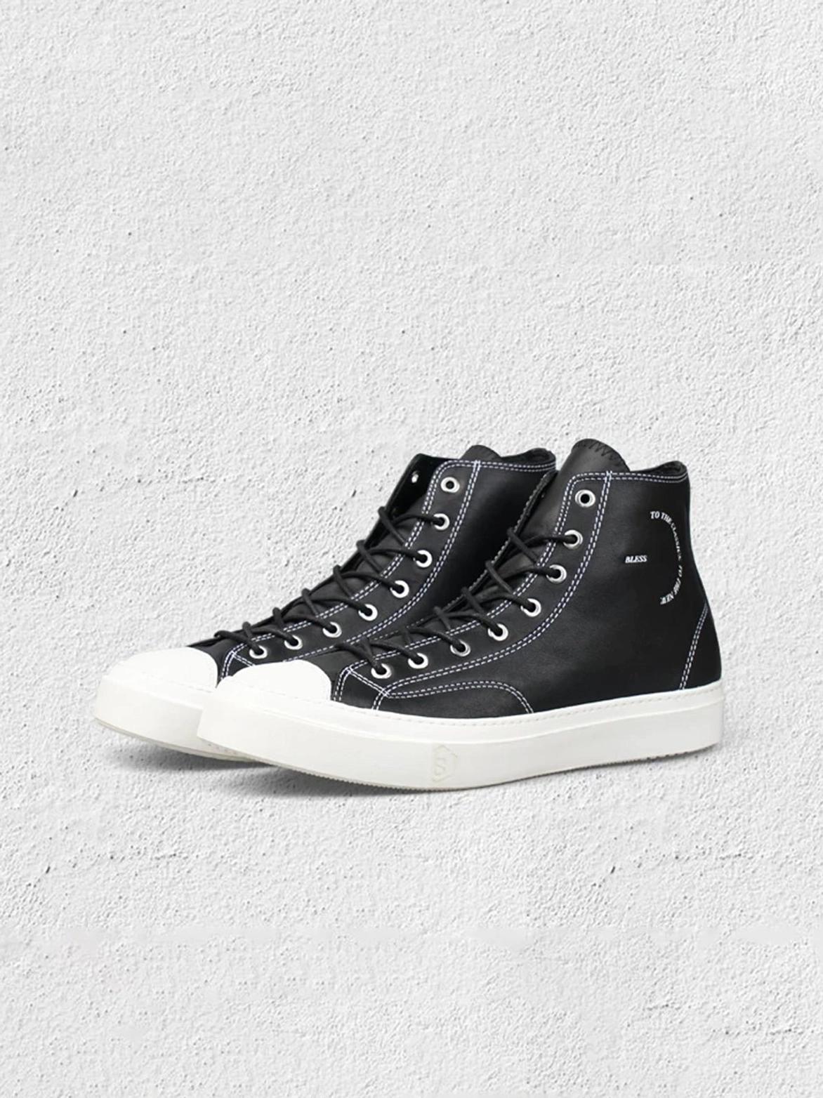 Pair of black contrast-stitch high-top platform sneakers displayed against a textured concrete background with white soles and printed heel details