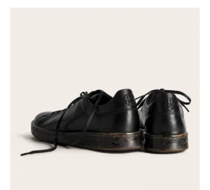 Pair of black leather derby sneakers shown from the rear three-quarter angle with vintage worn-tone rubber soles and structured heel panels