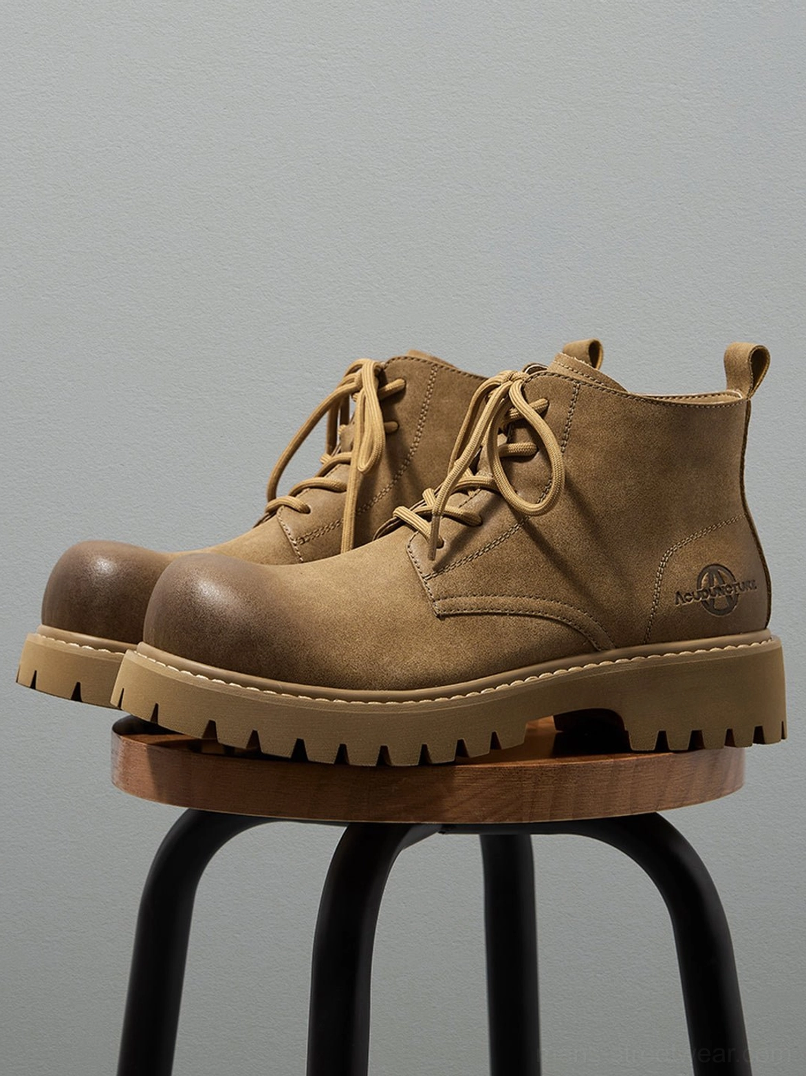 Pair of tan distressed work boots displayed on a wooden stool with rugged round-toe streetwear styling