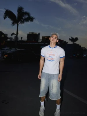 Full-body male streetwear outfit wearing a light grey graphic ringer T-shirt with blue trim, embellished denim shorts, white socks, and casual sneakers