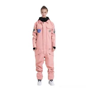 Pastel pink astronaut-inspired waterproof one-piece ski suit on female model with embroidered mission patches, front closure, and black ribbed cuffs