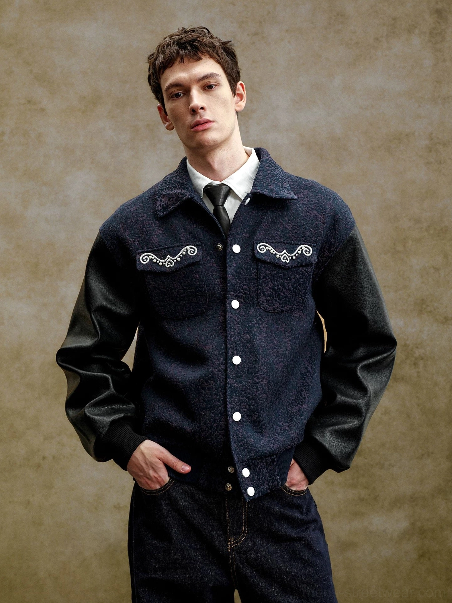 Front view of a pearl embellished navy varsity jacket with black faux leather contrast sleeves, snap buttons, textured body, and ornate chest details