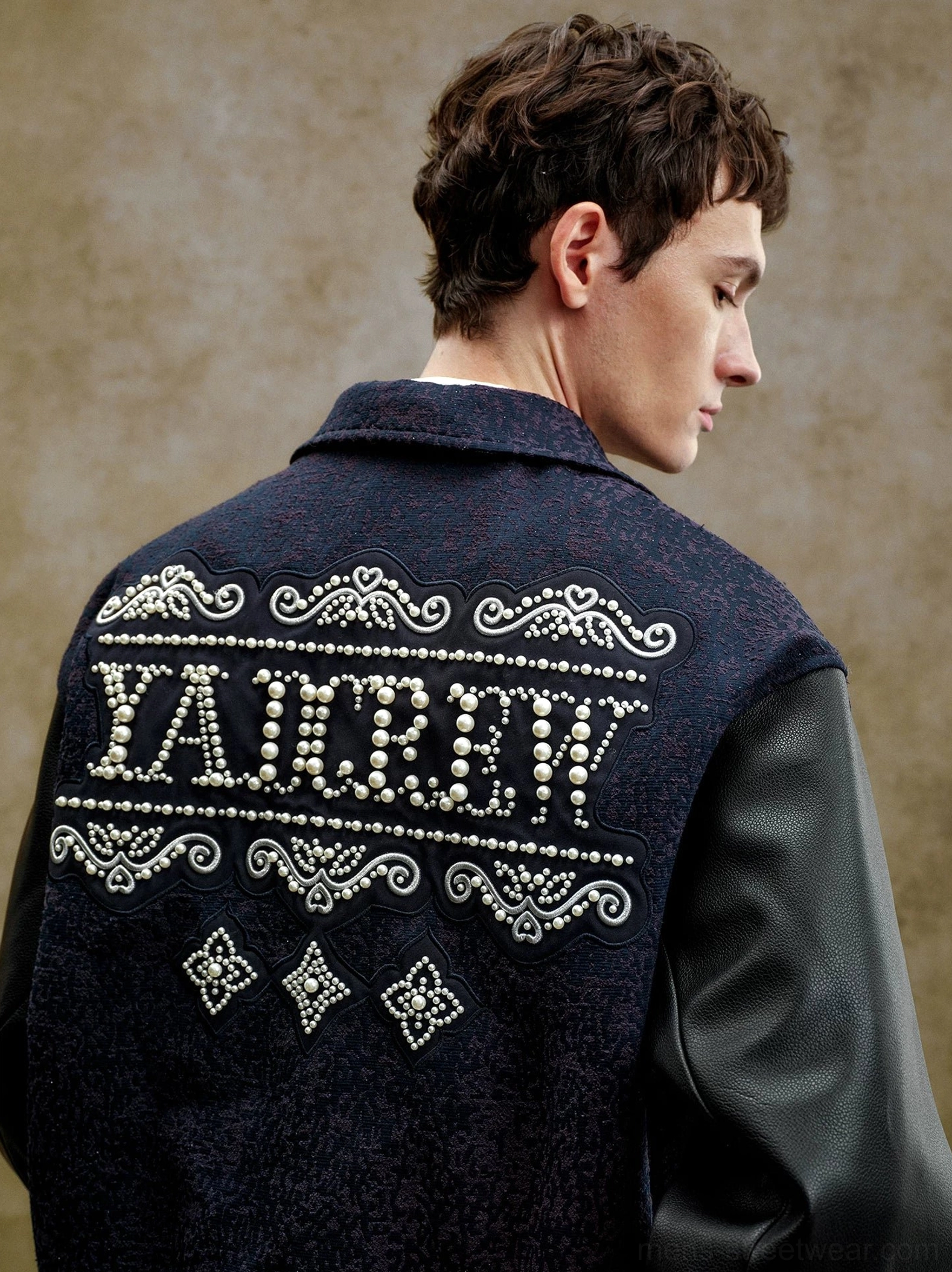 Back view of a pearl embellished varsity jacket showing oversized decorative lettering, ornate scroll motifs, diamond details, and black contrast sleeves