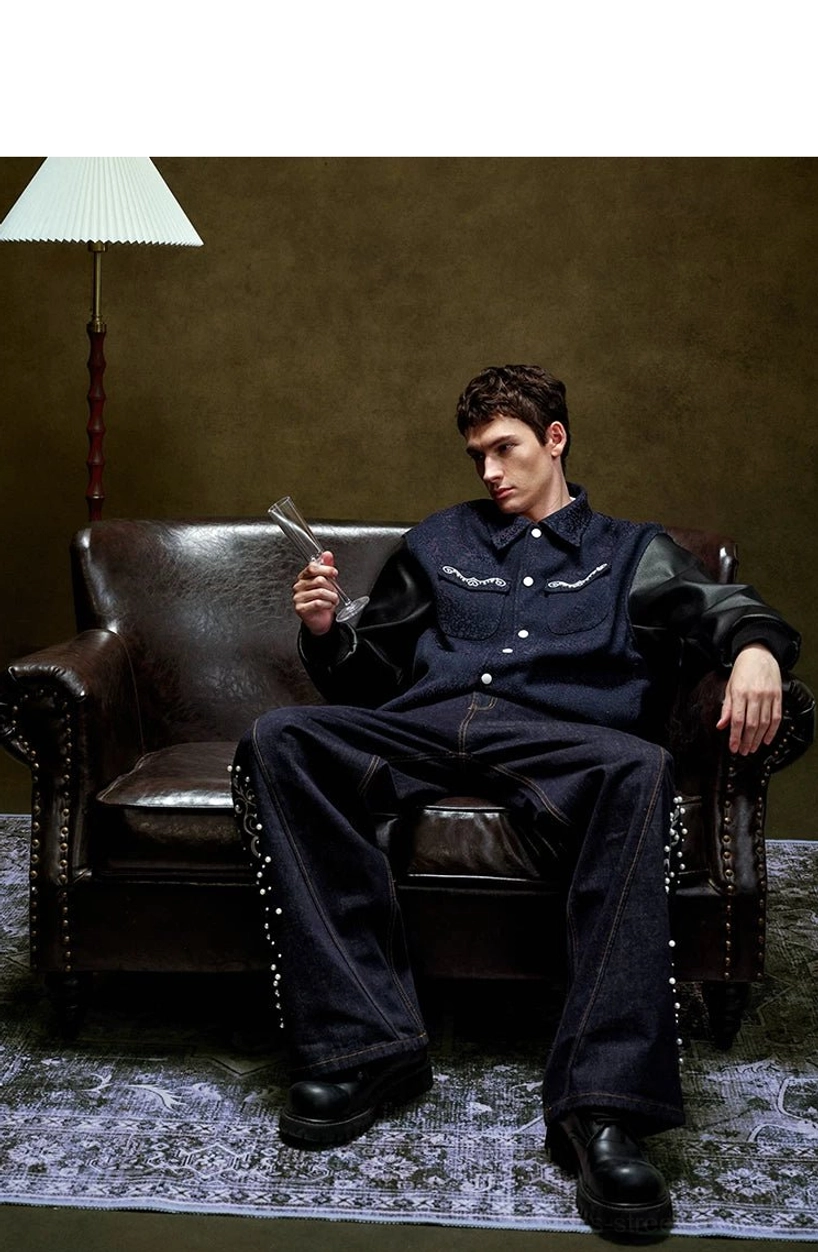 Full body seated view of a relaxed pearl embellished varsity jacket styled with embroidered denim pants in a vintage studio setting