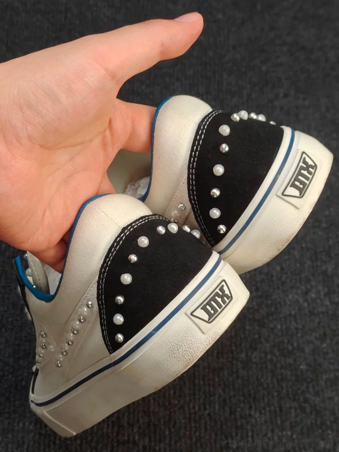 Close-up heel view of a pearl-studded low-top skate sneaker showing the black heel cap, silver embellishments, white upper, and blue trim detail