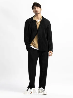 Black pleated straight-leg trousers styled with a blazer, khaki top, white undershirt, and casual sneakers in a minimalist streetwear studio look