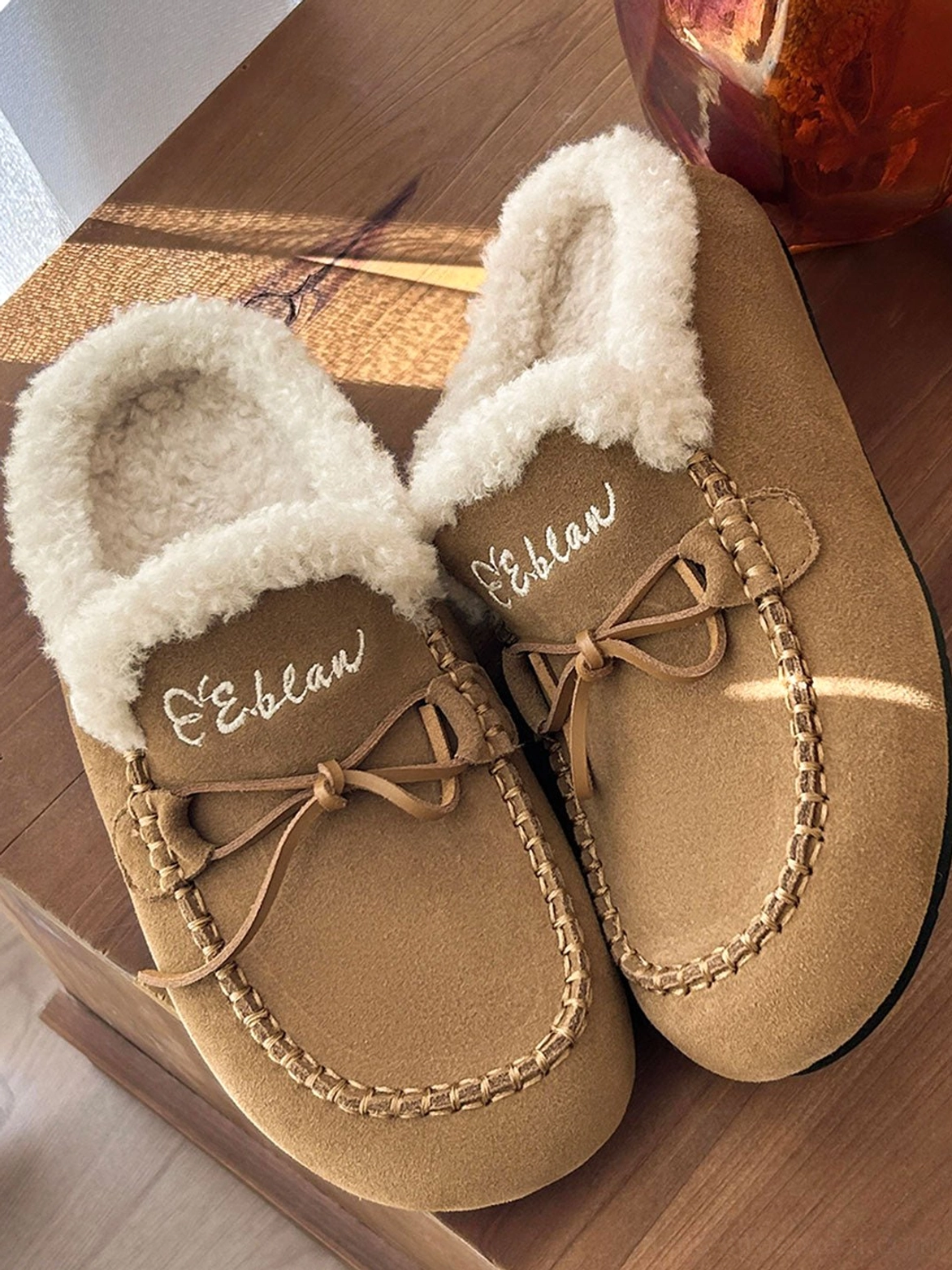 Close-up pair of tan plush-lined moccasin slippers showing stitched toe seams, fleece collar, and lace bow