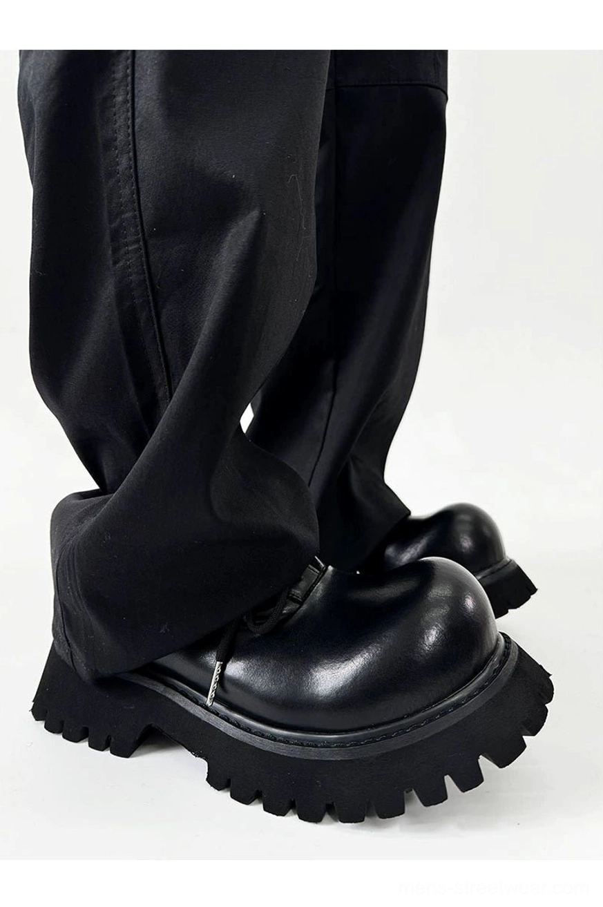Polished black Derby shoes with chunky lug soles worn with loose fit black trousers in studio setting