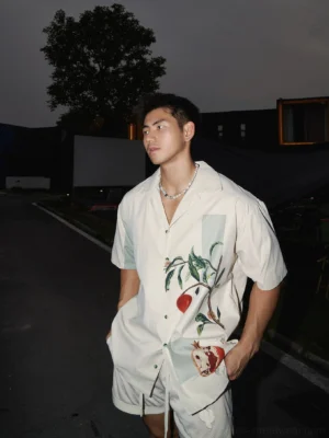 Three-quarter view of a relaxed beige pomegranate botanical print camp shirt styled with matching summer shorts on a male model outdoors at night