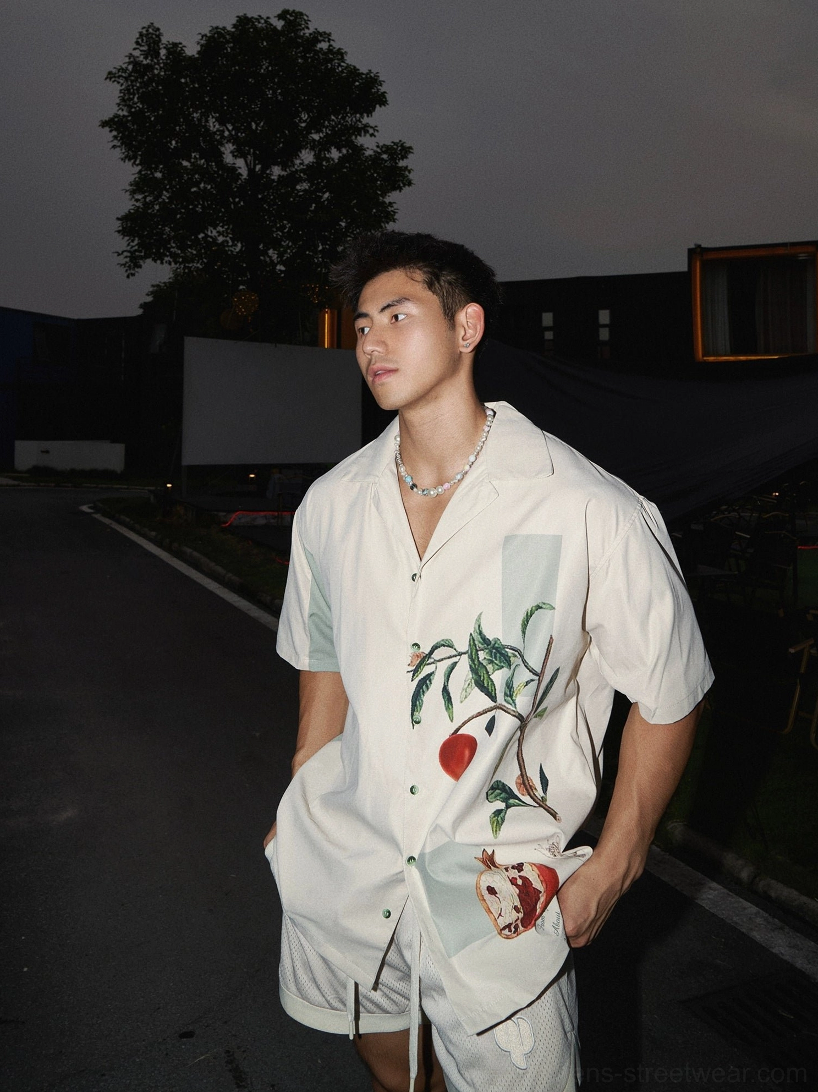 Three-quarter view of a relaxed beige pomegranate botanical print camp shirt styled with matching summer shorts on a male model outdoors at night