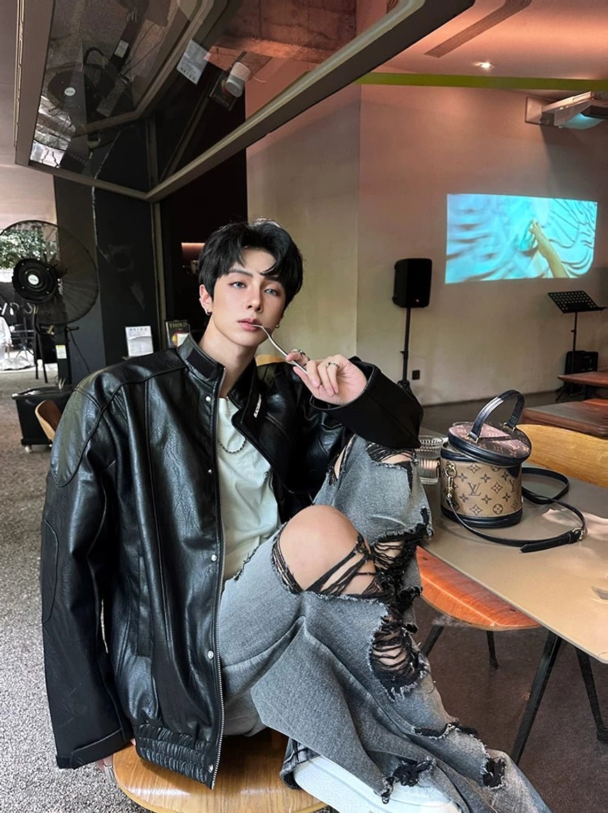 Male model seated in distressed gray wide-leg jeans with ripped thigh and knee details, black leather jacket, and white inner tee