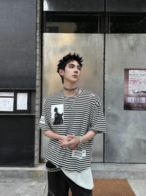 Full front view of a relaxed black and white striped streetwear T-shirt with vintage figure patches, sleeve details, punk accessories, and faux layered hem