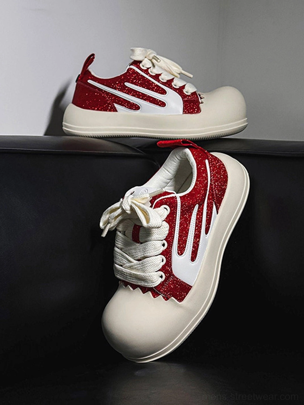Red glitter stripe chunky platform sneakers with oversized white round toe caps displayed on a black leather sofa
