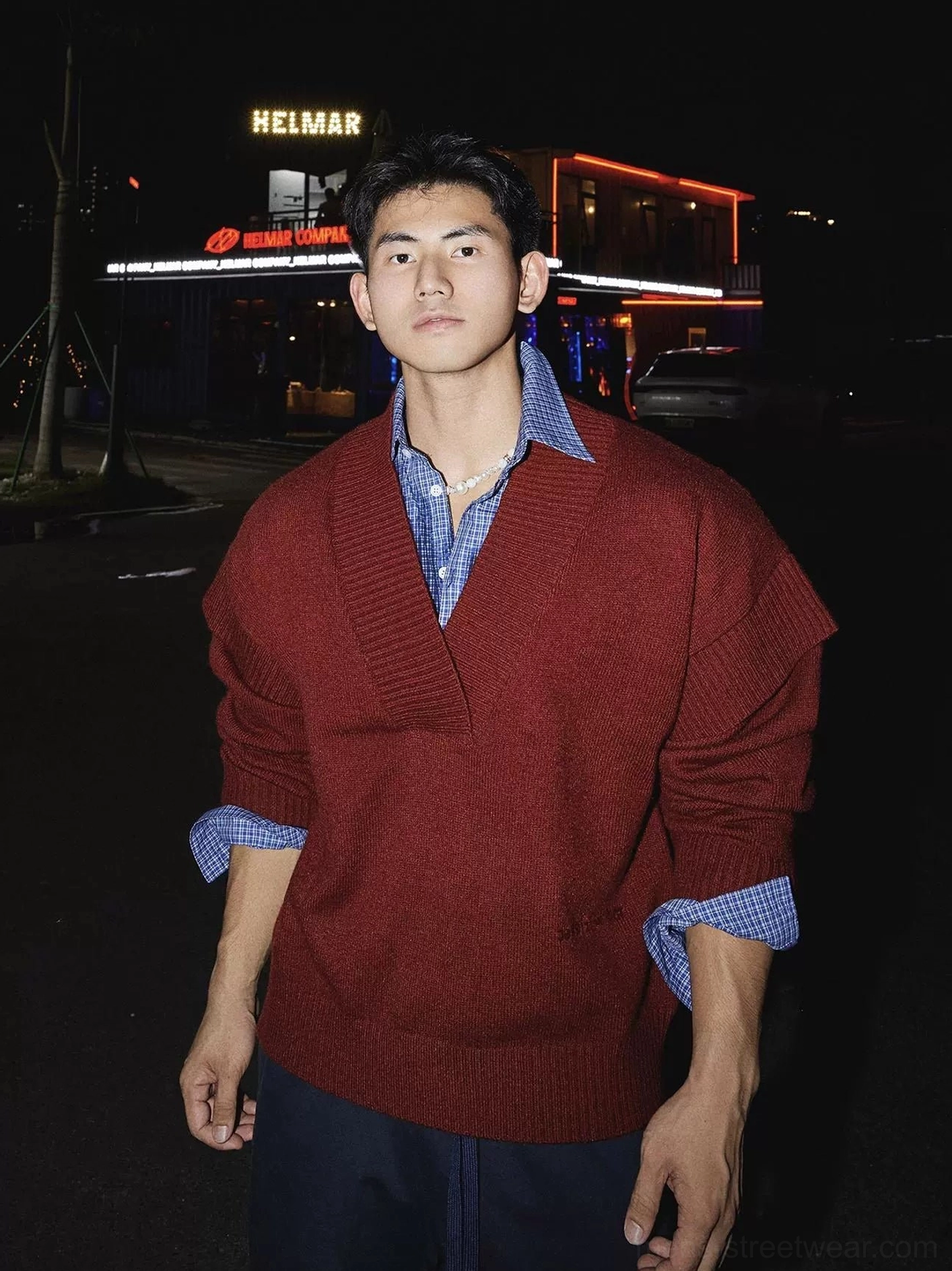 Red oversized deep V-neck knit sweater layered over a blue checked shirt on a male model in a nighttime streetwear setting