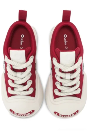 Front overhead view of red and white canvas low-top sneakers with burgundy upper, thick white rubber toe caps, woven laces, and playful smile graphic for Korean streetwear