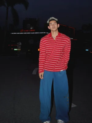 Full body front view of a model wearing a red and white striped long sleeve rugby polo shirt with loose blue wide leg trousers in a modern streetwear setting