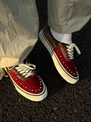 Reddish-brown studded low-top streetwear sneakers worn with beige loose cargo pants and white crew socks on an asphalt pavement background