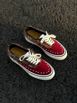 Reddish-brown studded suede low-top sneakers displayed on asphalt with burgundy suede toe caps, fuzzy brown heel panels, silver studs, white laces, and striped rubber soles