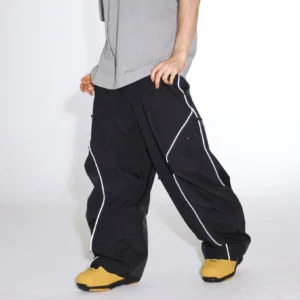 Black reflective wide-leg snow pants worn with yellow snow boots, showing oversized baggy snowboard fit, contrast piping, and side utility pockets
