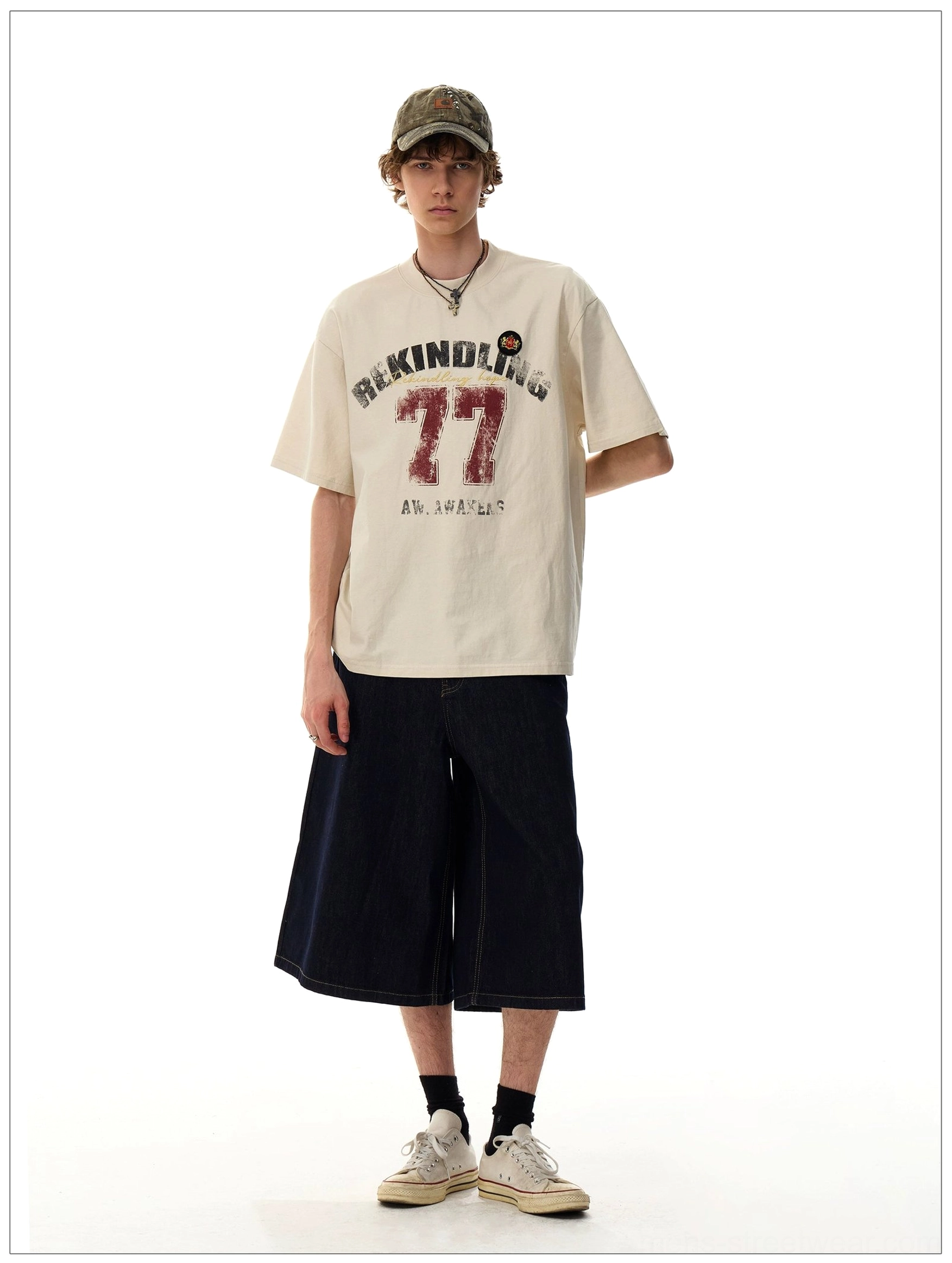 Side front view of the Apricot Rekindling 77 vintage varsity graphic T-shirt styled with denim shorts and a camouflage cap