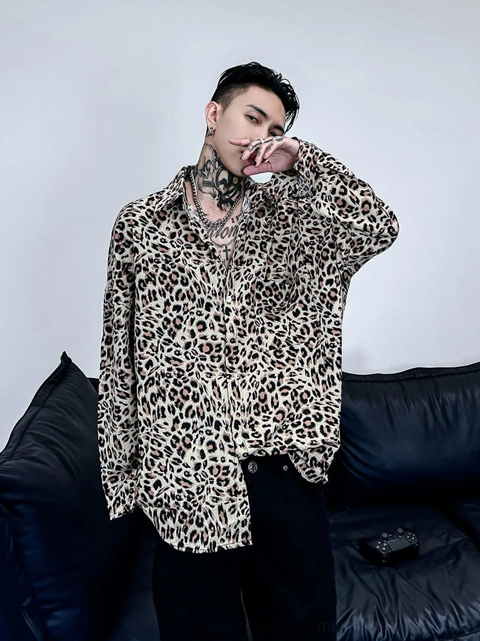 Three-quarter front view of a tattooed male model wearing a relaxed oversized leopard print satin shirt with long sleeves in a modern streetwear setting
