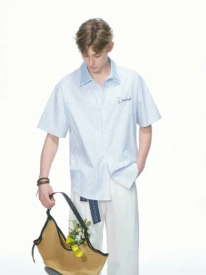 Blue and white pinstripe short sleeve contrast collar shirt styled with loose off white pants and a floral woven tote bag in a minimalist studio