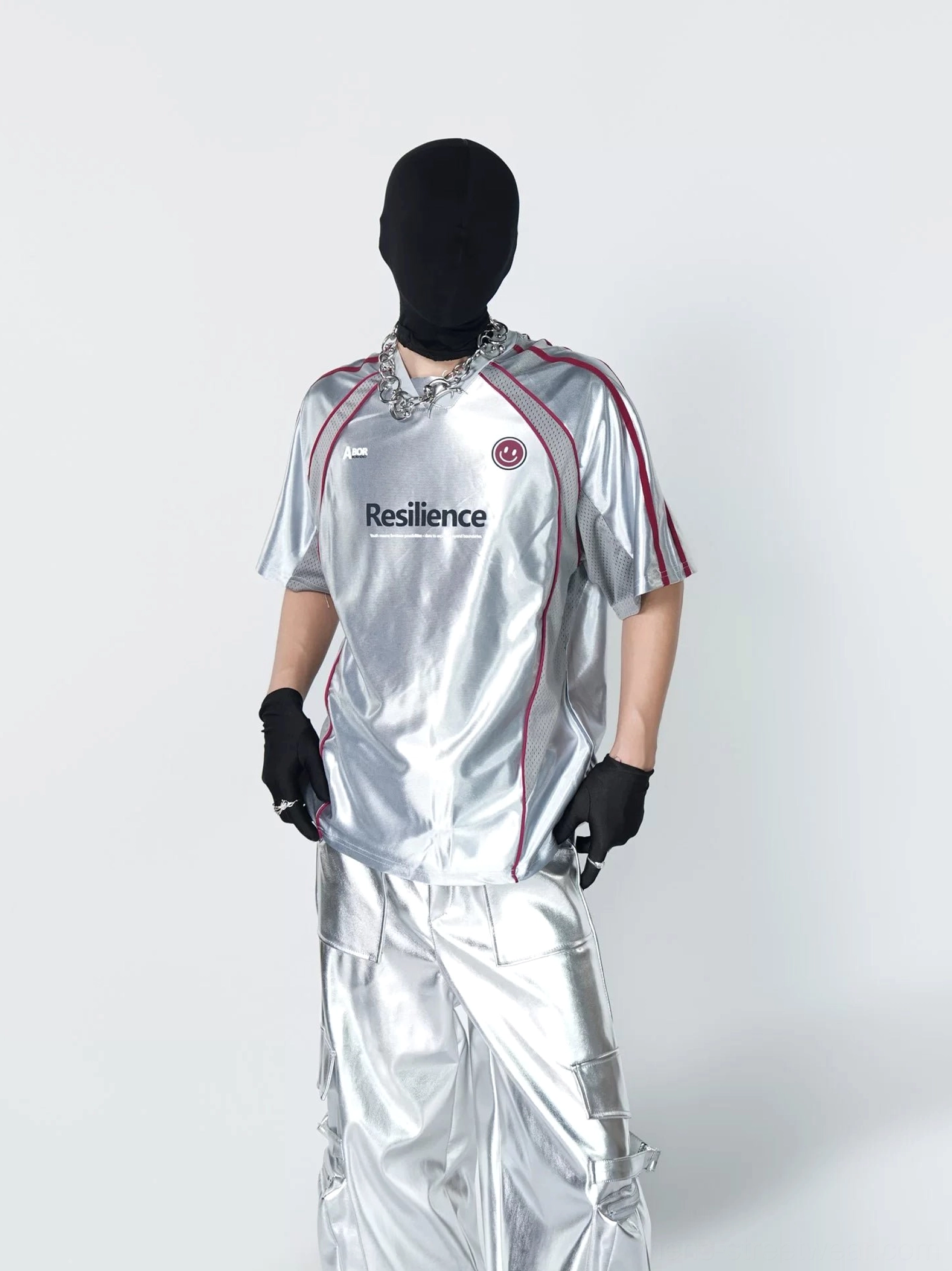 Close front view of metallic silver Resilience jersey top showing chest graphic, circular emblem, red seam piping, and techwear details