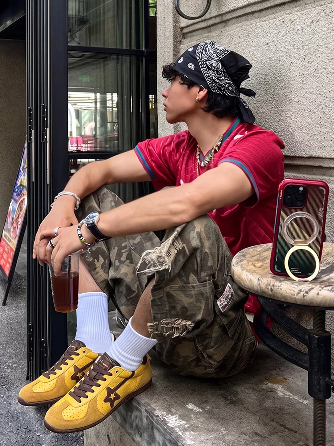 Yellow brown low-profile suede trainers styled with red tee and camouflage cargo pants at an outdoor urban cafe