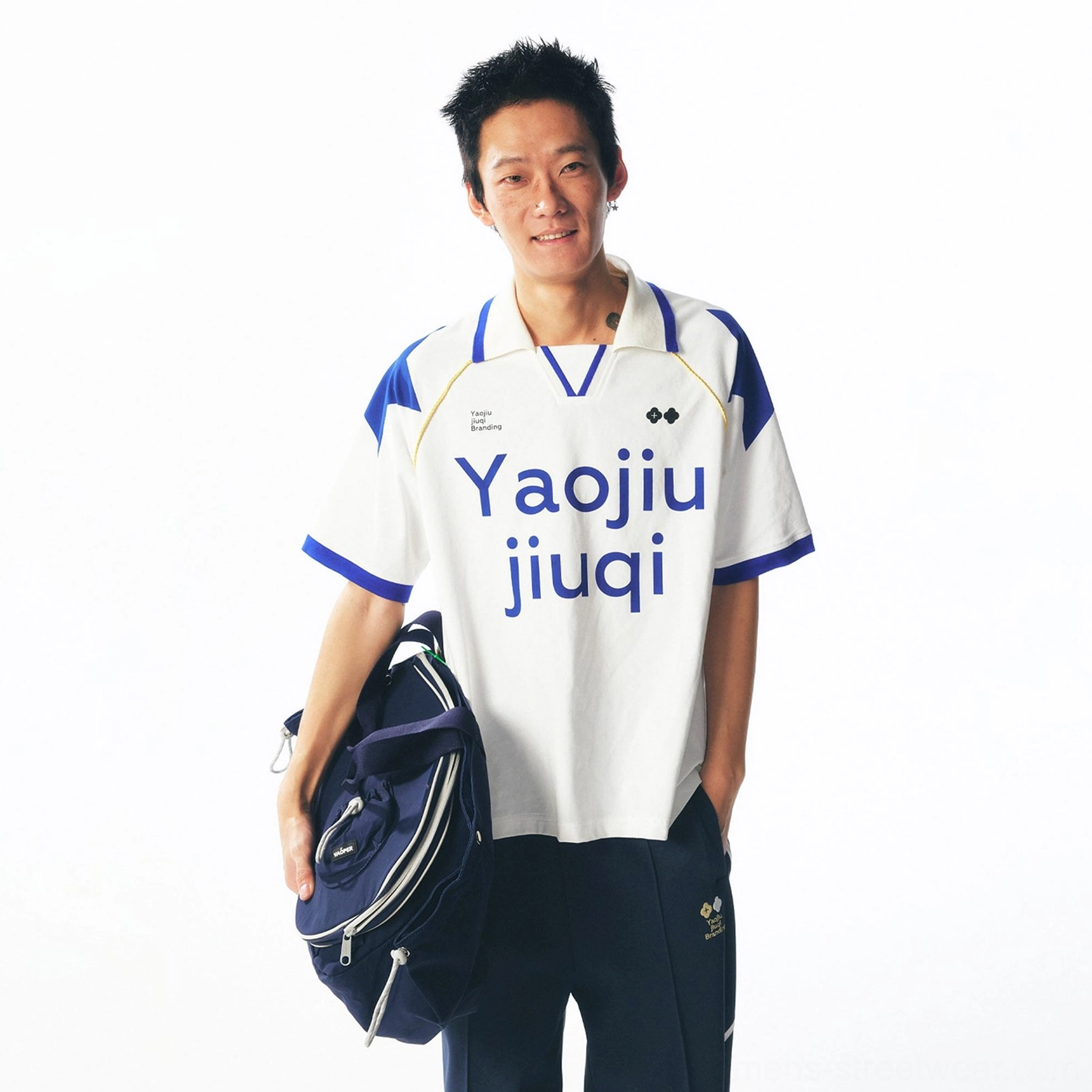 Front view of a retro white and blue contrast-panel V-neck football jersey with Yaojiu jiuqi typography, styled with navy sport pants and a dark duffel bag