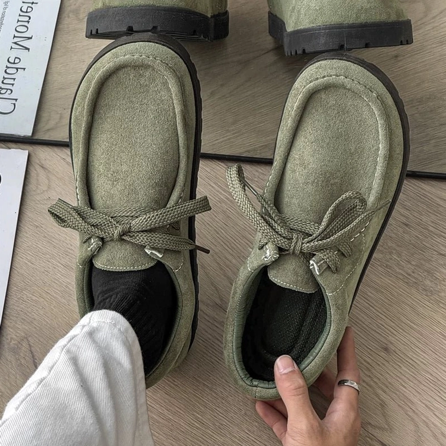 Sage green square-toe suede lace-up streetwear shoes with matching green laces, black ridged sole, black socks, and grey trousers