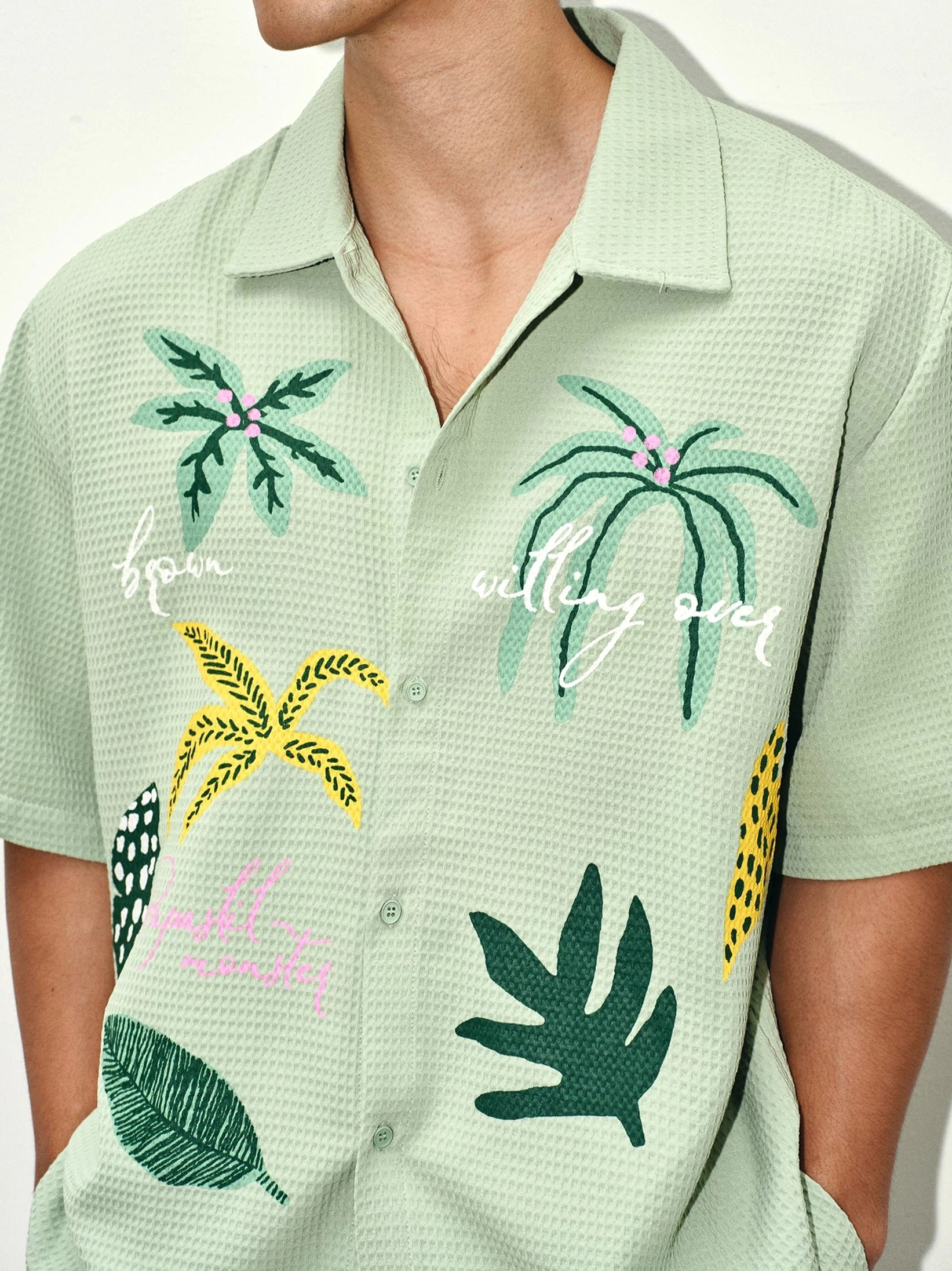 Close-up of sage green waffle texture camp shirt with button front palm leaf embroidery and handwritten tropical graphics