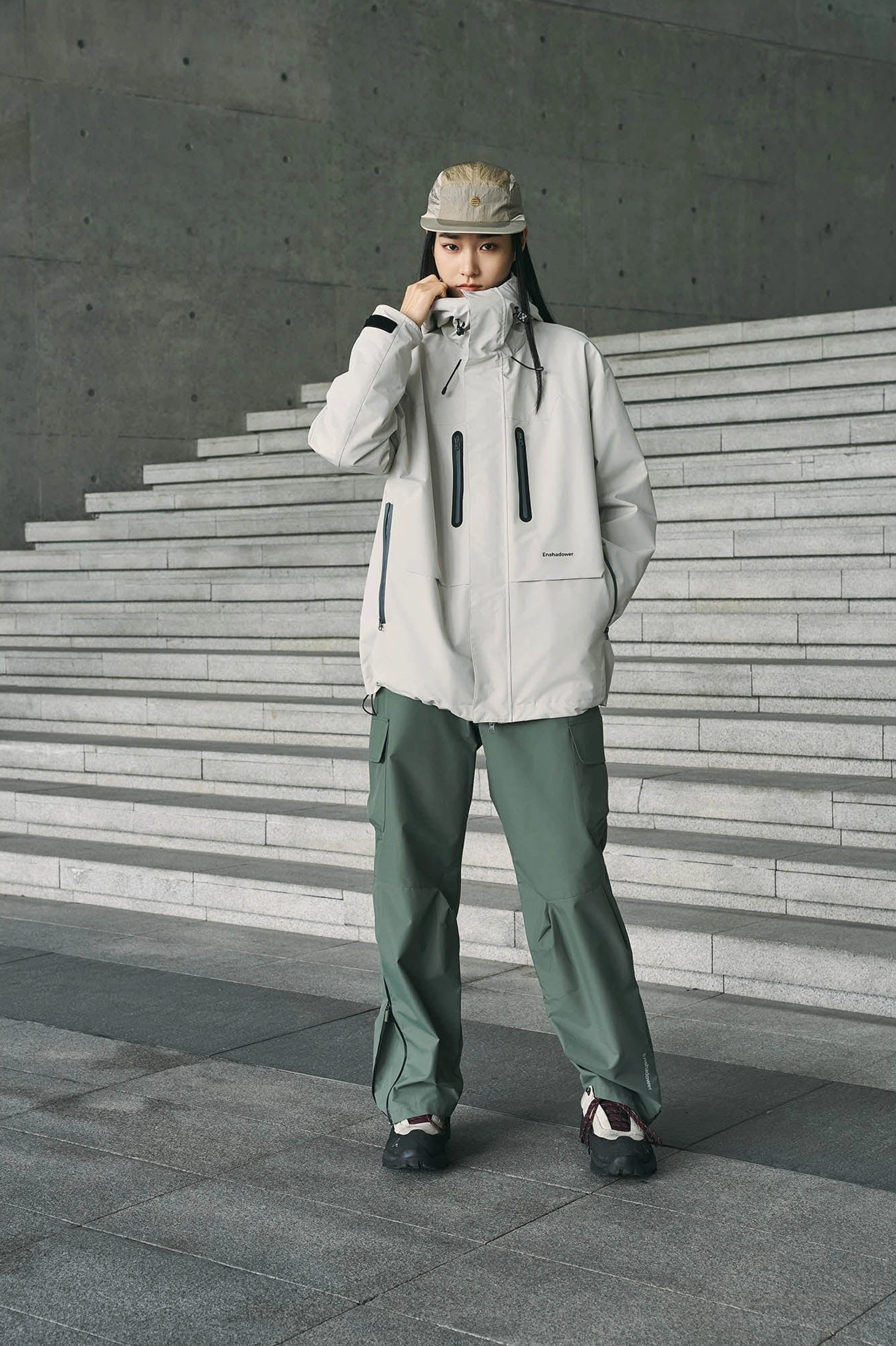 Sage green waterproof techwear pants styled with a light gray outdoor jacket, bucket hat, and casual sneakers