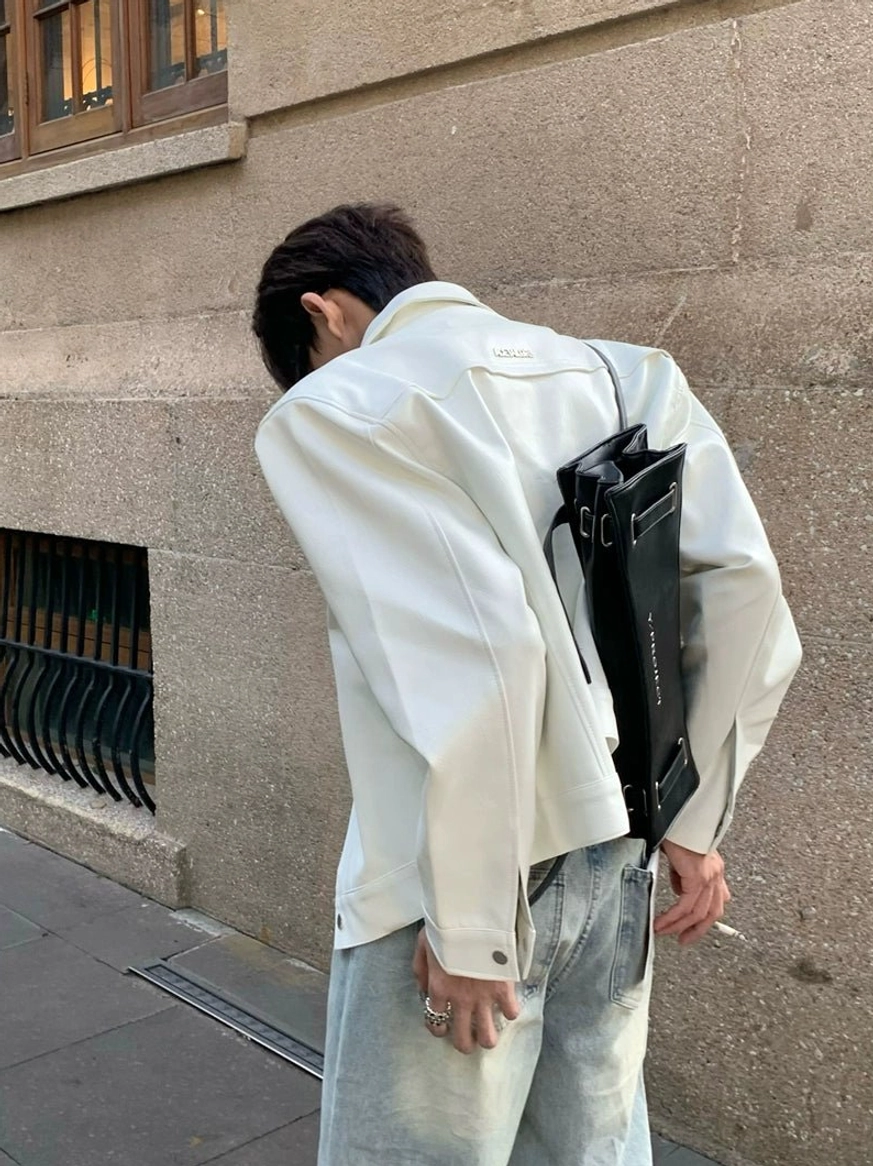 Off-white sculpted wide-shoulder cropped denim jacket shown from the rear three-quarter view on a male model in light blue jeans and a black crossbody bag on an urban stone street
