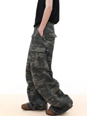 Side profile of a model wearing dark shadow camouflage wide leg cargo pants with oversized side pockets, black sleeveless streetwear top, and brown work boots
