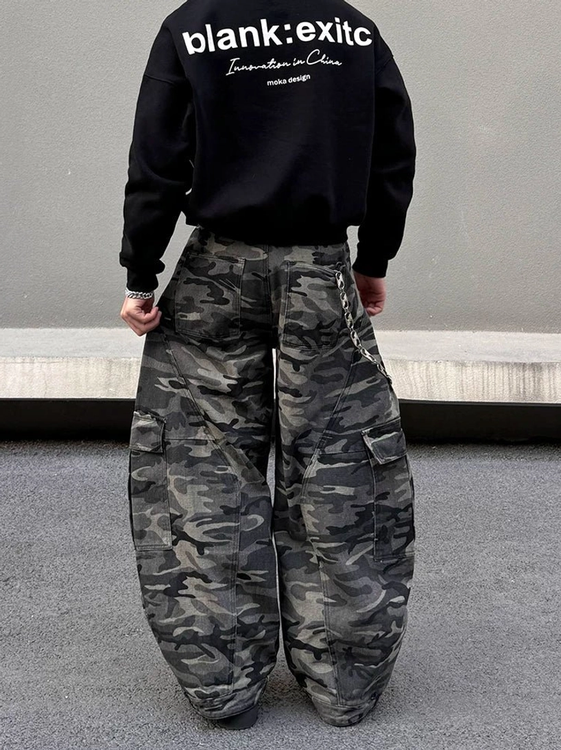 Back view of model wearing dark grey camouflage balloon cargo pants with oversized wide legs, side pockets, waist chain, and black streetwear sweatshirt