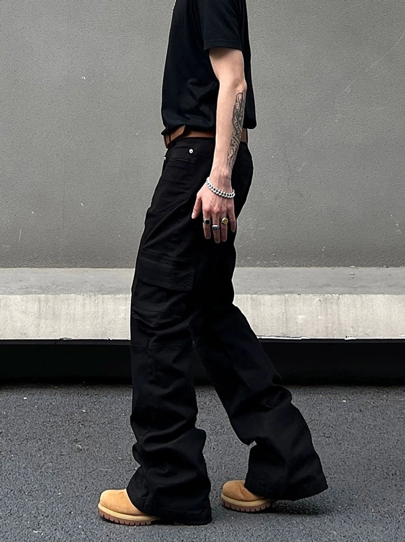 Side walking view of black cargo flared pants showing stacked hems, utility pockets, brown leather belt, and tan work boots