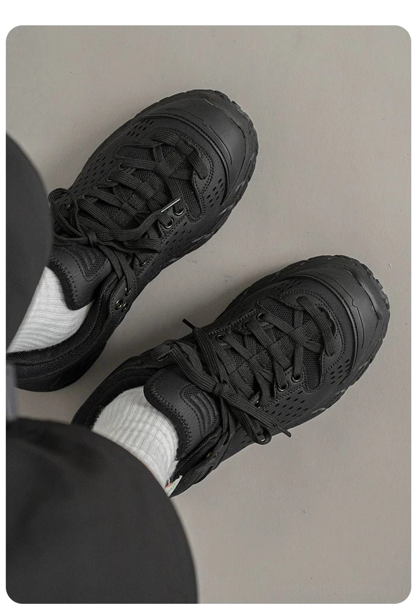 Overhead flat lay of black low-top utility sneakers with perforated upper, thick treaded sole, and lace-up closure beside black clothing