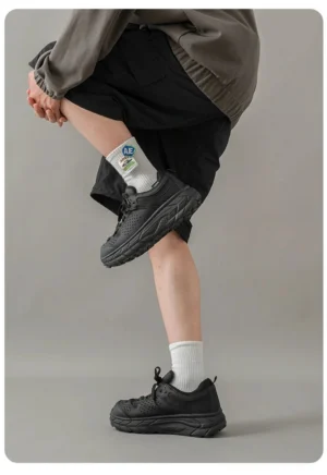 Male model wearing matte black chunky utility trainers with a rugged anti-slip trail sole, gray sweatshirt, black shorts, and white socks in a minimalist studio