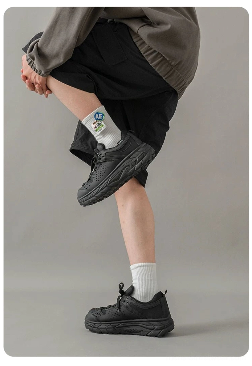 Male model wearing matte black chunky utility trainers with a rugged anti-slip trail sole, gray sweatshirt, black shorts, and white socks in a minimalist studio