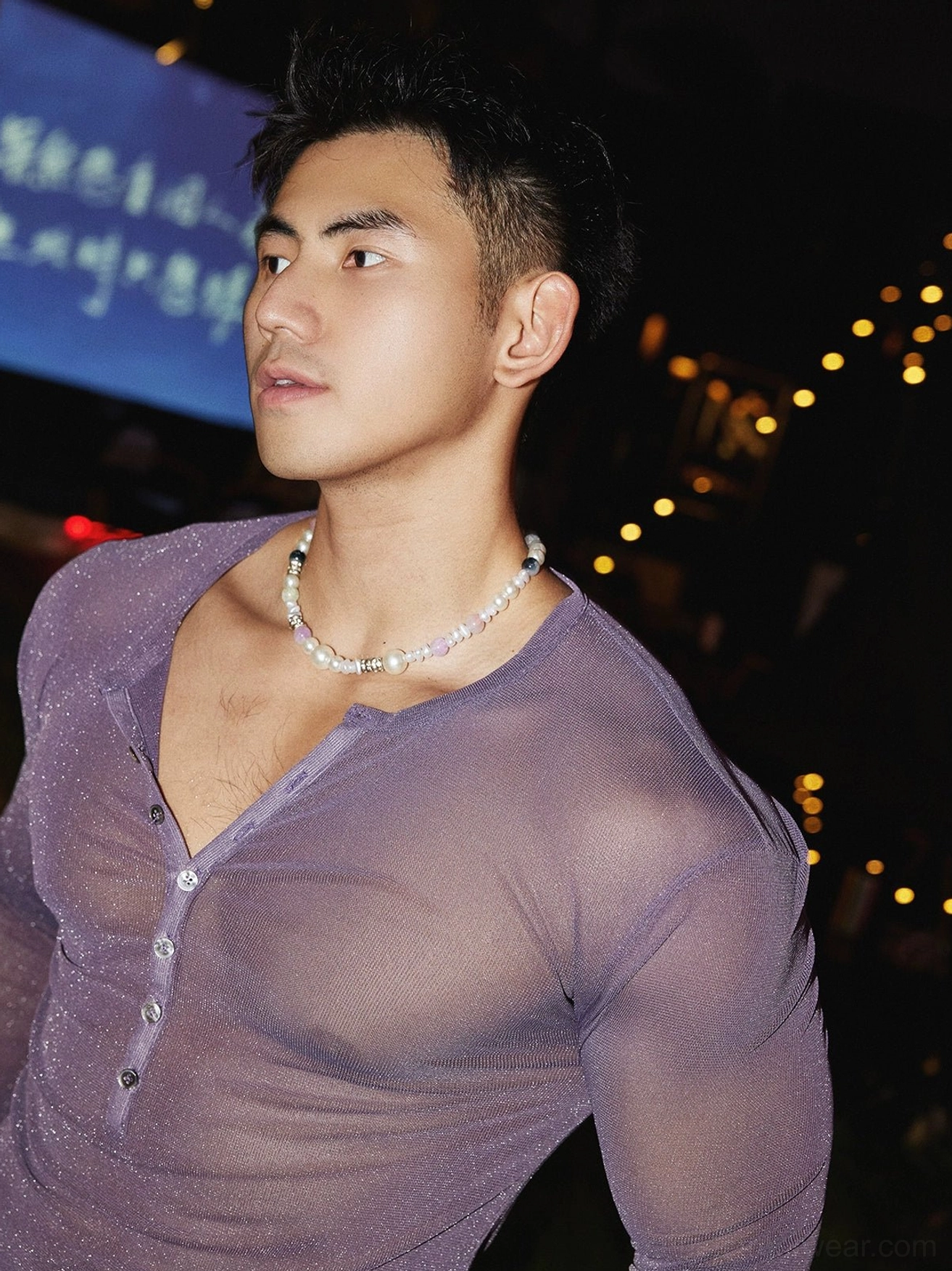 Side close-up of a male model wearing a light purple sheer sparkly Henley mesh top with pearl necklace under decorative night lights