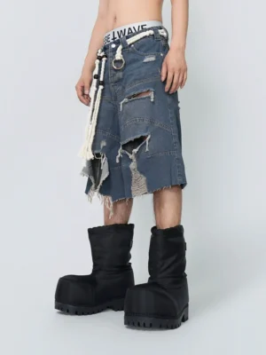 Front three quarter view of wide shredded indigo denim shorts with distressed rips, raw asymmetrical hem, thick white rope belt, metal ring hardware, and black platform boots