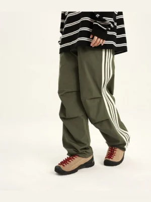 Army green side striped athletic pants with three white stripes, loose straight-leg fit, and knee pleats styled with retro sneakers