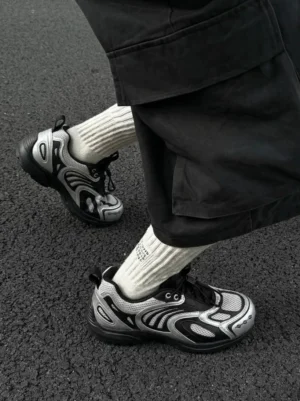 Silver black mesh runner sneakers worn with black cargo pants and cream ribbed socks on an asphalt road, showing the chunky anti-slip rubber outsole and futuristic streetwear design