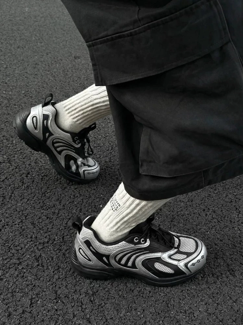 Silver black mesh runner sneakers worn with black cargo pants and cream ribbed socks on an asphalt road, showing the chunky anti-slip rubber outsole and futuristic streetwear design