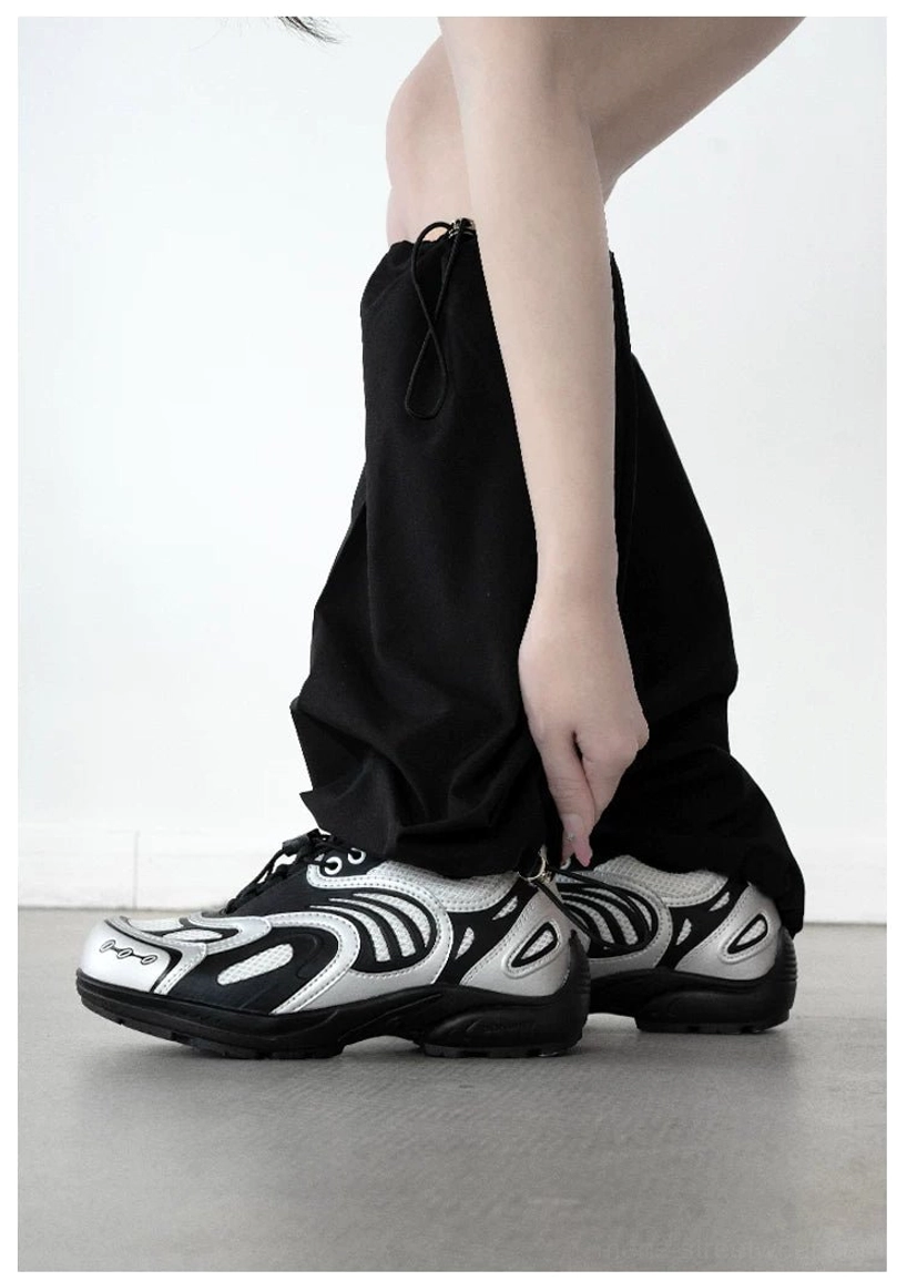 Rear view of silver black techwear sneakers styled with drawstring cargo pants on a concrete floor, highlighting symmetrical silver textures and retro futuristic streetwear detailing