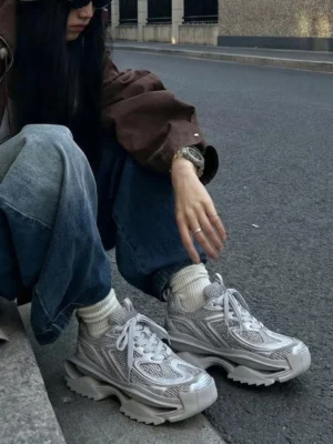 Silver chunky mesh streetwear sneakers worn with white crew socks in an urban roadside setting, showing the oversized sculpted outsole and Korean fashion styling