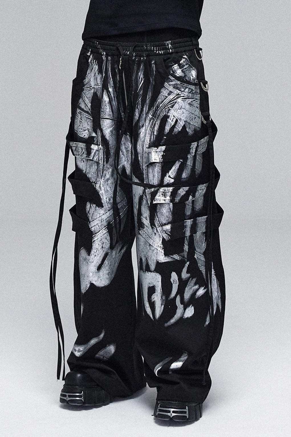 Low-angle detail of silver graffiti cargo pants featuring layered utility pockets, full abstract print, and industrial side drawstrings